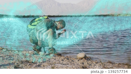 Crouching hiker scooping water from lakeshore at calm lake, with green backpack and hiking boots 139257734