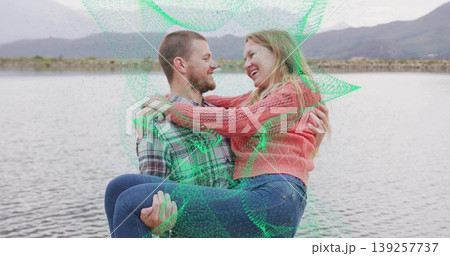 Man in plaid shirt carrying woman wearing pink sweater on rocky shore, with calm lake, mountains 139257737