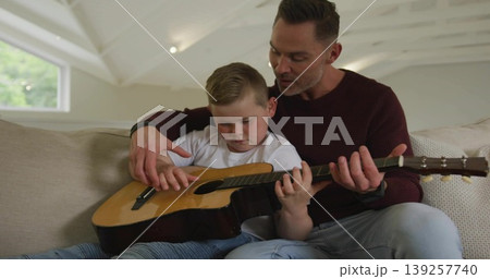 Father in maroon shirt guiding son playing acoustic guitar on sofa in living room, with focus 139257740