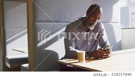 Holding smartphone, senior Black man wearing shirt sitting at modern lounge table with coffee cup Holding smartphone, senior Black man wearing shirt sitting at modern lounge table with coffee cup 139257946