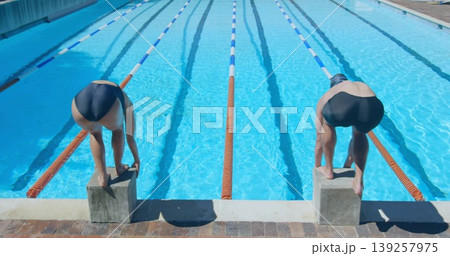 Swimmers wearing caps crouching on starting blocks in outdoor lap pool, with lane ropes and goggles 139257975