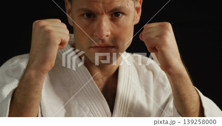 Raising fists mid adult male holding defensive guard stance in studio, with white martial arts gi Raising fists mid adult male holding defensive guard stance in studio, with white martial arts gi 139258000