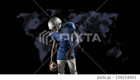 Holding brown football, helmet-wearing player standing in sports studio, lit world map Holding brown football, helmet-wearing player standing in sports studio, lit world map 139258001