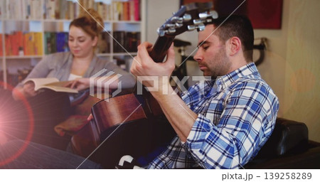 Strumming man playing acoustic guitar on armchair in living room, bookshelf and bicycle, copy space 139258289