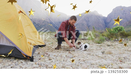 Squatting camper wearing red plaid shirt on rocky campsite, engaging terrier dog near yellow tent Squatting camper wearing red plaid shirt on rocky campsite, engaging terrier dog near yellow tent 139258300