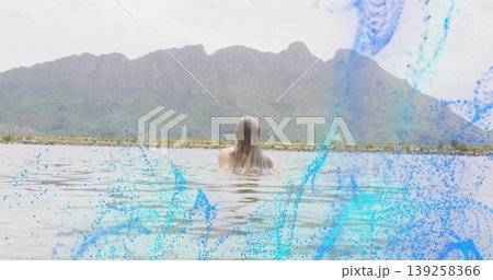 Woman in swimsuit standing in lake facing mountains under clouds, with digital splash graphics 139258366