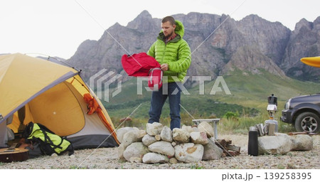 Camper wearing green jacket standing at mountain camp, with yellow tent, firepit, stove and thermos Camper wearing green jacket standing at mountain camp, with yellow tent, firepit, stove and thermos 139258395