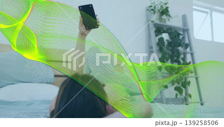 Scrolling smartphone, woman propping on pillow in bedroom with potted plant on shelf near window Scrolling smartphone, woman propping on pillow in bedroom with potted plant on shelf near window 139258506