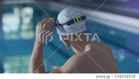 Adult male swimmer adjusting swim cap while holding goggles in aquatic facility with marked lanes Adult male swimmer adjusting swim cap while holding goggles in aquatic facility with marked lanes 139258576
