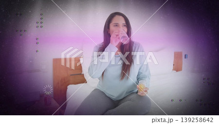 Sipping Indian female in shirt and leggings on bed, with translucent pink cup and clear container 139258642