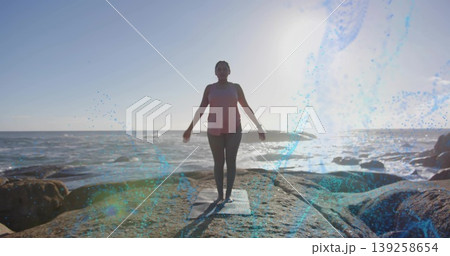 Woman in coral top practicing yoga balancing on rocky shoreline, with yoga mat and smartwatch 139258654
