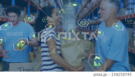 Volunteer wearing light blue Tshirt handing paper bag to colleague by warehouse boxes and eco icons 139258745