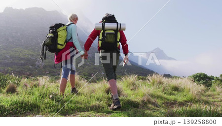 Walking mature couple ascending grassy mountain slope, carrying backpacks with rolled mats and map 139258800