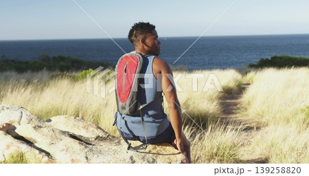 Man wearing blue tank top sitting, gazing at ocean on dune rock, with hydration backpack 139258820
