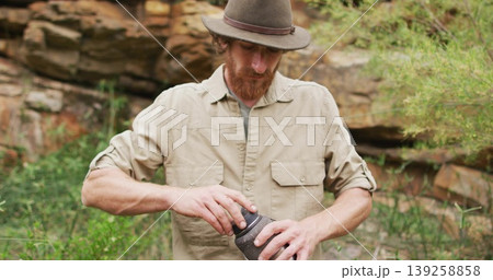Twisting open container, bearded man wearing felt hat and khaki shirt outdoors at rock outcrop Twisting open container, bearded man wearing felt hat and khaki shirt outdoors at rock outcrop 139258858