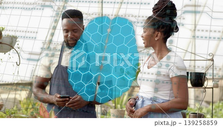 Nursery worker wearing apron showing smartphone app to assistant in greenhouse, with potted plants Nursery worker wearing apron showing smartphone app to assistant in greenhouse, with potted plants 139258859