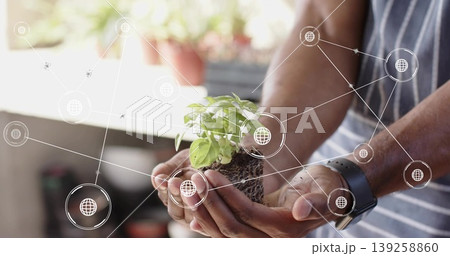 Man wearing apron holding seedling in soil by garden window, smartwatch, digital globe overlays Man wearing apron holding seedling in soil by garden window, smartwatch, digital globe overlays 139258860