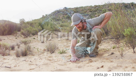Explorer wearing baseball cap, over-ear headphones, touching sand in shrubland with digital swirl Explorer wearing baseball cap, over-ear headphones, touching sand in shrubland with digital swirl 139258867