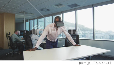 Leaning Black male wearing VR headset on white table in modern office, with dual monitors, mouse 139258931
