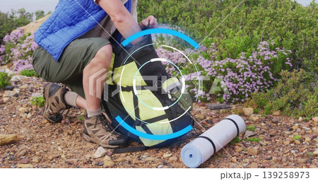 Unpacking male hiker wearing blue vest organizing backpack on hillside, with rolled sleeping mat 139258973