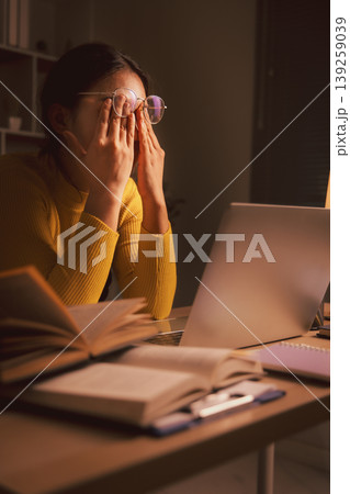 Young Asian woman in glasses rubbing tired eyes while using laptop in cozy home office at night, showing screen fatigue, study burnout, remote work stress, eyestrain and late productivity. 139259039