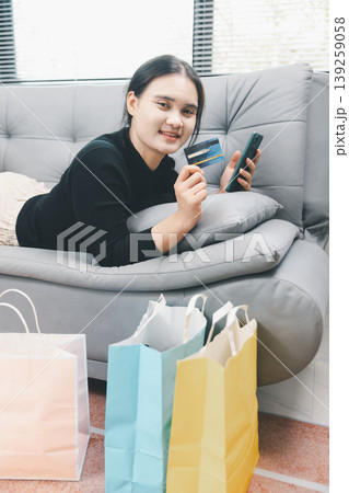 Young Asian woman lying on sofa holding smartphone and credit card with shopping bags at home, online shopping, mobile payment, ecommerce lifestyle, cashless consumer and retail concept 139259058