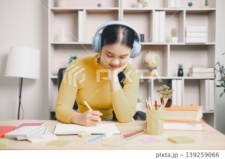Young Asian woman student in headphones writing study notes at home desk with books, pencils and sticky notes. Focused learning, exam preparation, concentration, education and lifestyle concept. Young Asian woman student in headphones writing study notes at home desk with books, pencils and sticky notes. Focused learning, exam preparation, concentration, education and lifestyle concept. 139259066