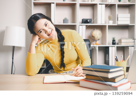 Young Asian woman studying online at home desk, writing notes with earphones and books beside laptop. Smiling student, remote learning, virtual class, education and motivation concept. Young Asian woman studying online at home desk, writing notes with earphones and books beside laptop. Smiling student, remote learning, virtual class, education and motivation concept. 139259094