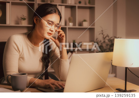 Young Asian woman in glasses working on laptop in cozy home office at night, showing deep focus, remote work, online study, problem solving, concentration and calm evening productivity. Young Asian woman in glasses working on laptop in cozy home office at night, showing deep focus, remote work, online study, problem solving, concentration and calm evening productivity. 139259135