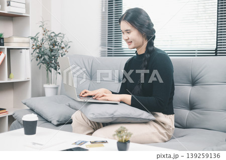 Young Asian woman using laptop on sofa at home for online shopping, browsing ecommerce website with credit cards on table, digital payment, cashless purchase and consumer lifestyle concept 139259136