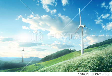 Two wind turbines rotate on green hills. The landscape features a clear blue sky filled with clouds. The sun shines bright, highlighting the green fields and trees nearby. Two wind turbines rotate on green hills. The landscape features a clear blue sky filled with clouds. The sun shines bright, highlighting the green fields and trees nearby. 139259327