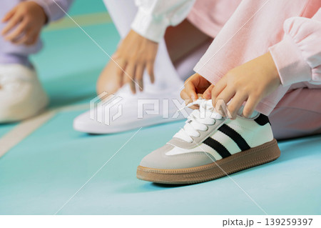 Two children are getting ready for a sports practice. One child is tying the shoelaces on white and black shoes while sitting on a blue gym floor in the afternoon. Two children are getting ready for a sports practice. One child is tying the shoelaces on white and black shoes while sitting on a blue gym floor in the afternoon. 139259397