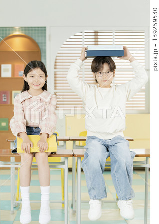 Two children are sitting at their desks in a classroom. One child holds a book above their head while the other smiles and holds a notebook. Two children are sitting at their desks in a classroom. One child holds a book above their head while the other smiles and holds a notebook. 139259530