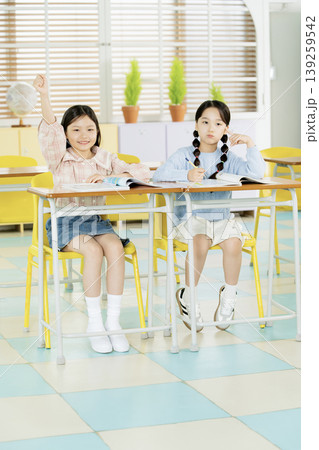 In a bright classroom, two girls are seated at desks. One girl raises her hand, indicating she wants to answer a question, while the other watches attentively. Sunlight comes through the windows. 139259542