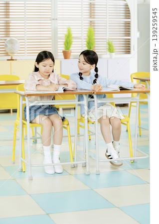 Two girls are seated at their desks in a classroom. They are looking at their books while surrounded by bright yellow chairs and plants. This scene takes place during school time. Two girls are seated at their desks in a classroom. They are looking at their books while surrounded by bright yellow chairs and plants. This scene takes place during school time. 139259545