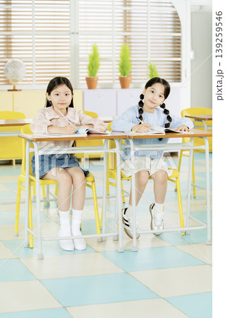 Two girls are seated at desks in a classroom. They wear casual clothes and sit in a colorful room with green plants. Other desks are visible behind them in the background. 139259546