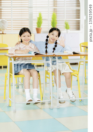 Two girls are sitting at a desk in a classroom. They are smiling and looking happy. One girl waves while the other sits closely and holds a book. The room is filled with light. Two girls are sitting at a desk in a classroom. They are smiling and looking happy. One girl waves while the other sits closely and holds a book. The room is filled with light. 139259549