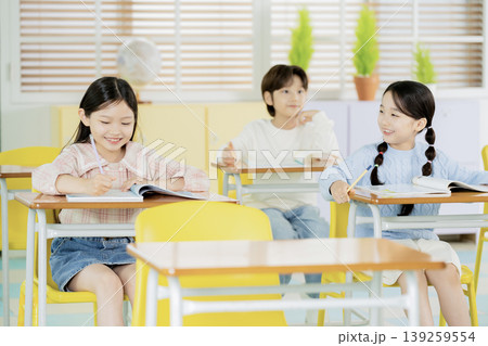 Three children sit at desks in a classroom. They are focused on their books and participate in learning. The room has windows and plants, creating a lively atmosphere. Three children sit at desks in a classroom. They are focused on their books and participate in learning. The room has windows and plants, creating a lively atmosphere. 139259554
