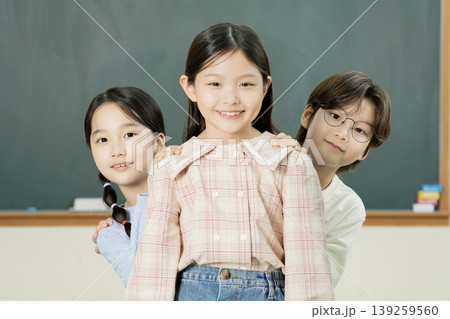Three children stand together in a classroom. One child is in front, while the others stand behind with smiles. They seem happy and engaged in a school activity during the day. Three children stand together in a classroom. One child is in front, while the others stand behind with smiles. They seem happy and engaged in a school activity during the day. 139259560