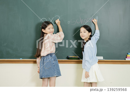 Two girls stand in a classroom writing on a chalkboard. They are preparing to start their school day. Each girl holds a piece of chalk in her hand. Two girls stand in a classroom writing on a chalkboard. They are preparing to start their school day. Each girl holds a piece of chalk in her hand. 139259568