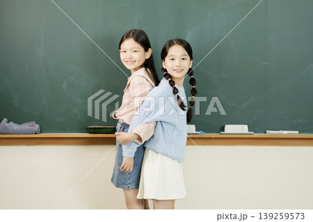 Two girls are standing back to back in a classroom. They are smiling and wearing casual clothes. The room has a green chalkboard and a desk in the background. Two girls are standing back to back in a classroom. They are smiling and wearing casual clothes. The room has a green chalkboard and a desk in the background. 139259573