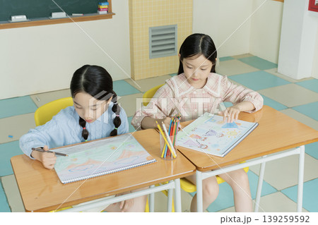 Two girls sit at their desks in a classroom and draw on their sketchbooks. They use colored pencils and focus on their artwork. The classroom has colorful tiles and chalkboard. Two girls sit at their desks in a classroom and draw on their sketchbooks. They use colored pencils and focus on their artwork. The classroom has colorful tiles and chalkboard. 139259592