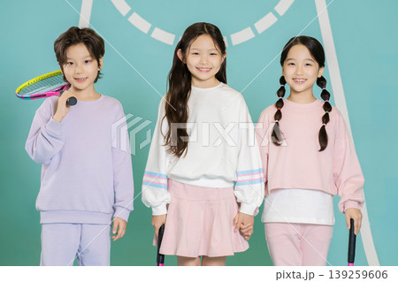 Three children stand together at a tennis court. They hold tennis rackets and wear matching outfits. They are smiling and look happy. The setting is bright and colorful, showing a playful mood. 139259606