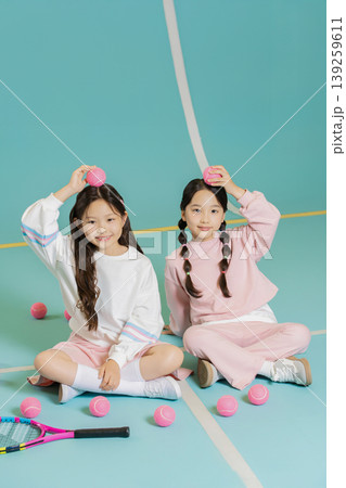 Two girls sit on a court and hold pink balls above their heads. They smile while surrounded by more pink balls and a tennis racket, enjoying their time together in a playful moment. 139259611