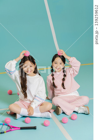 Two girls sit on a blue tennis court. They hold pink tennis balls on their heads and smile happily. The court is empty and bright. The activity looks fun and playful. Two girls sit on a blue tennis court. They hold pink tennis balls on their heads and smile happily. The court is empty and bright. The activity looks fun and playful. 139259612