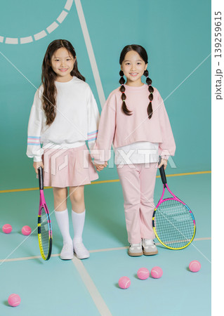 Two young girls hold tennis rackets while standing on a court. They are smiling at the camera, with pink tennis balls around them. They wear casual sports clothes and look happy. Two young girls hold tennis rackets while standing on a court. They are smiling at the camera, with pink tennis balls around them. They wear casual sports clothes and look happy. 139259615