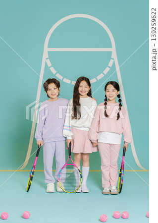 Three kids stand together holding tennis rackets in a bright blue space. They smile and wear sports clothes. Tennis balls are scattered on the ground around them. 139259622