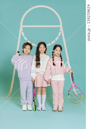 Three children stand together in a studio, each holding a tennis racket. They wear casual outfits in different colors and have a friendly pose. 139259624