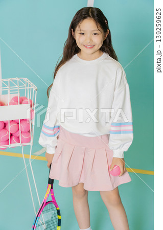 A young girl smiles while holding a tennis ball in her hand. She stands near a basket filled with pink tennis balls. The indoor court has blue walls and a simple design. A young girl smiles while holding a tennis ball in her hand. She stands near a basket filled with pink tennis balls. The indoor court has blue walls and a simple design. 139259625