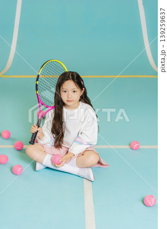 A young girl sits on the tennis court holding a racket with pink tennis balls around her. She focuses on her training while preparing for her next move in the sport. 139259637